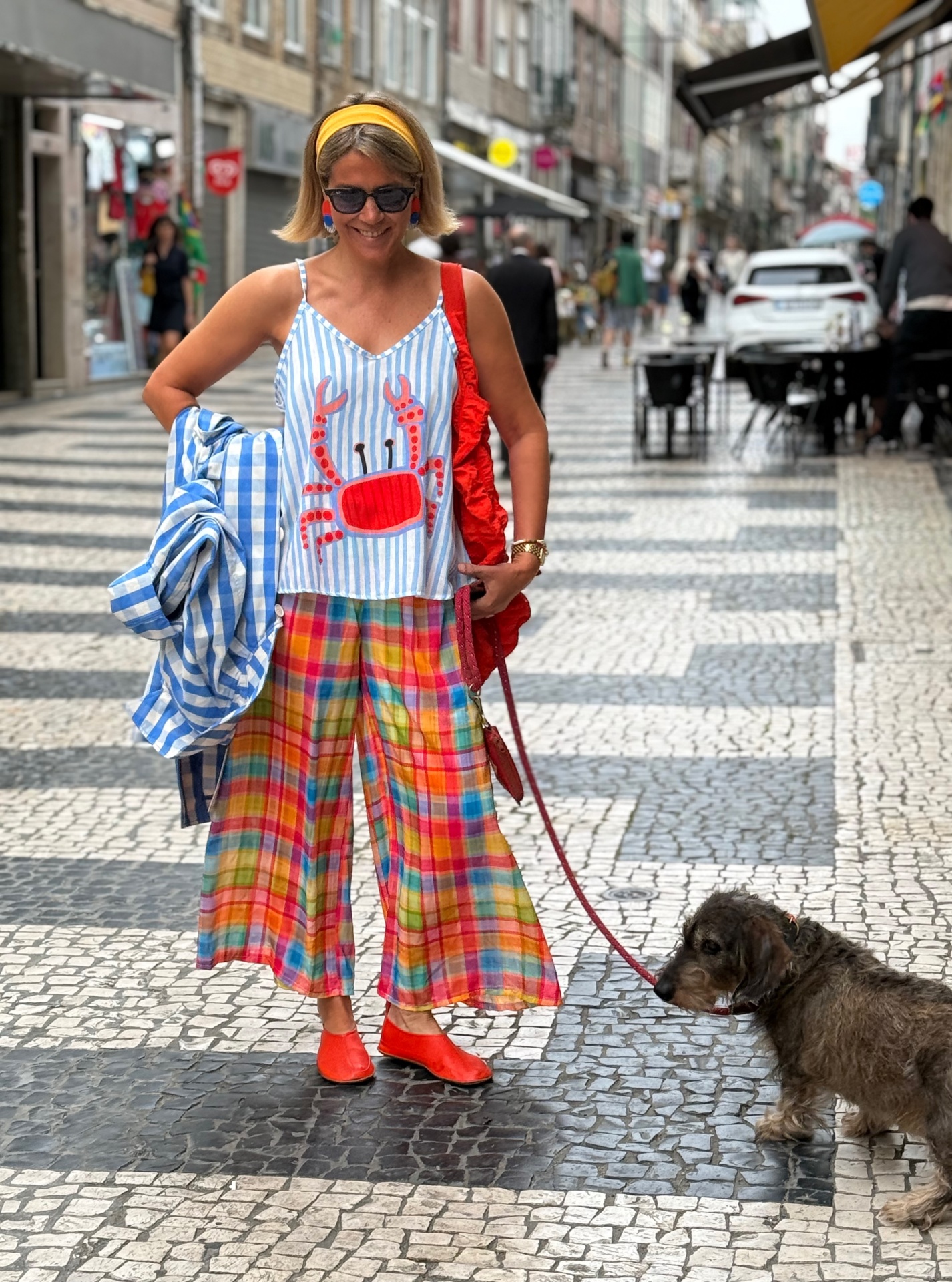 Mulher com top de alças com caranguejo, calças largas xadrez, sapatilhas vermelhas e lenço amarelo na cabeça numa rua calçada