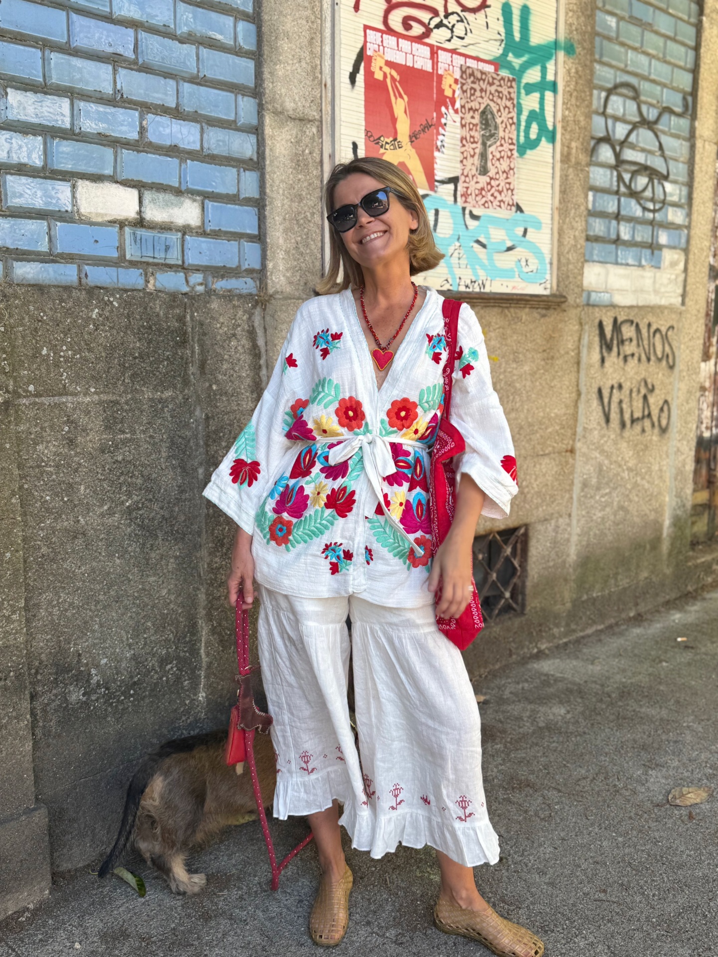 Mulher sorridente de camisa e calças brancas bordadas junto a parede com azulejos azuis e grafiti