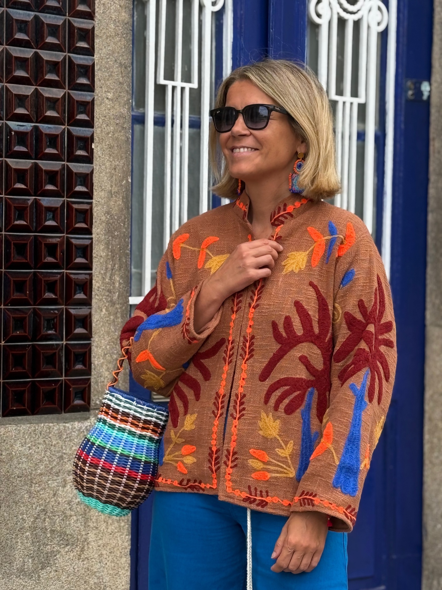 Mulher com casaco castanho bordado, calças azuis e saco de palha colorido a sorrir junto a porta azul