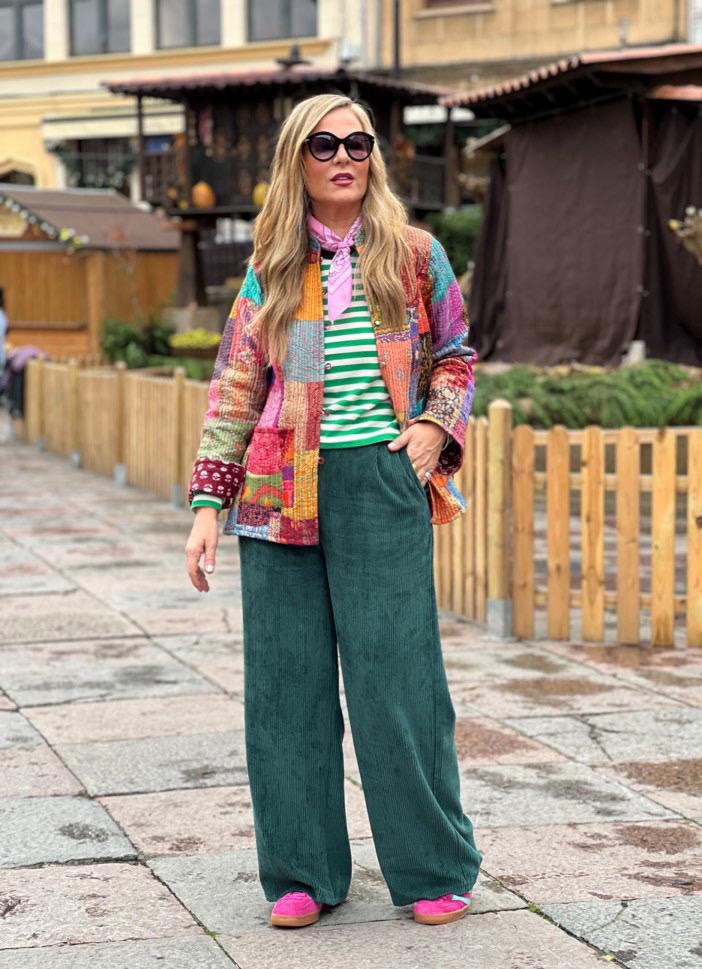 Mulher com casaco colorido de patchwork, t-shirt às riscas, calças verdes largas e lenço rosa, em zona exterior urbana.