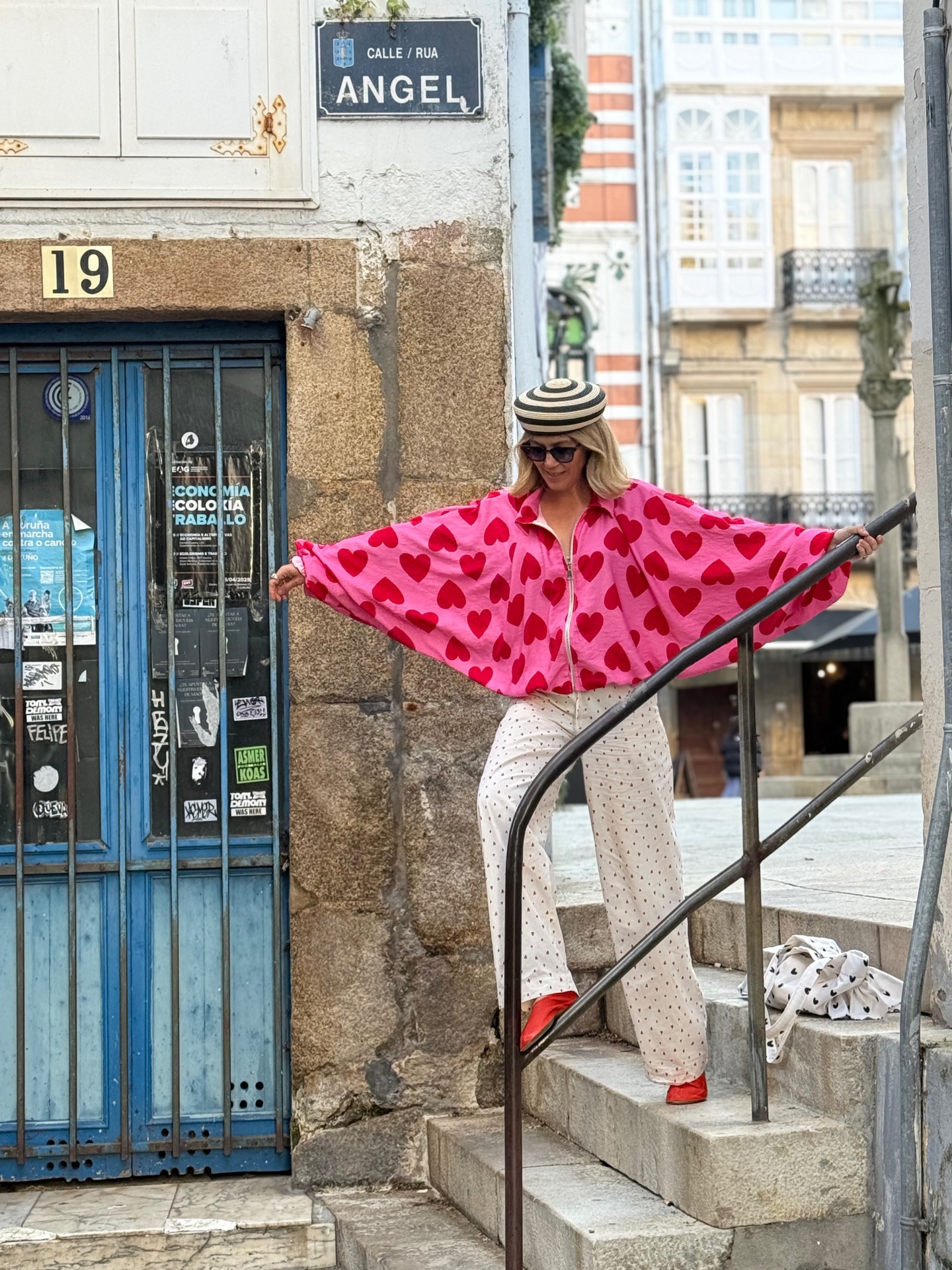 Mulher com casaco rosa com corações vermelhos, calças brancas com corações, sapatos vermelhos e chapéu listrado numa escadaria ao lado de porta azul.