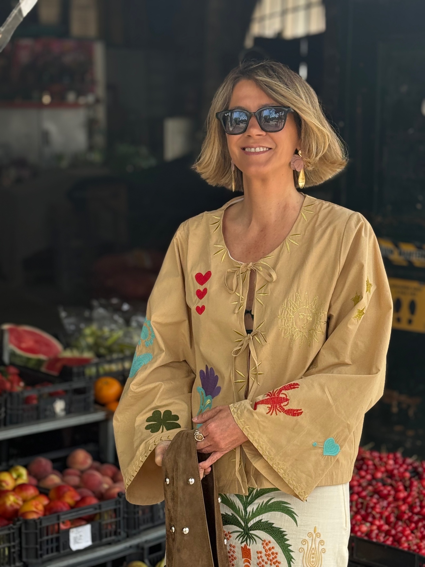 Blusa bege bordada com motivos coloridos num mercado com frutas e caixas ao fundo.