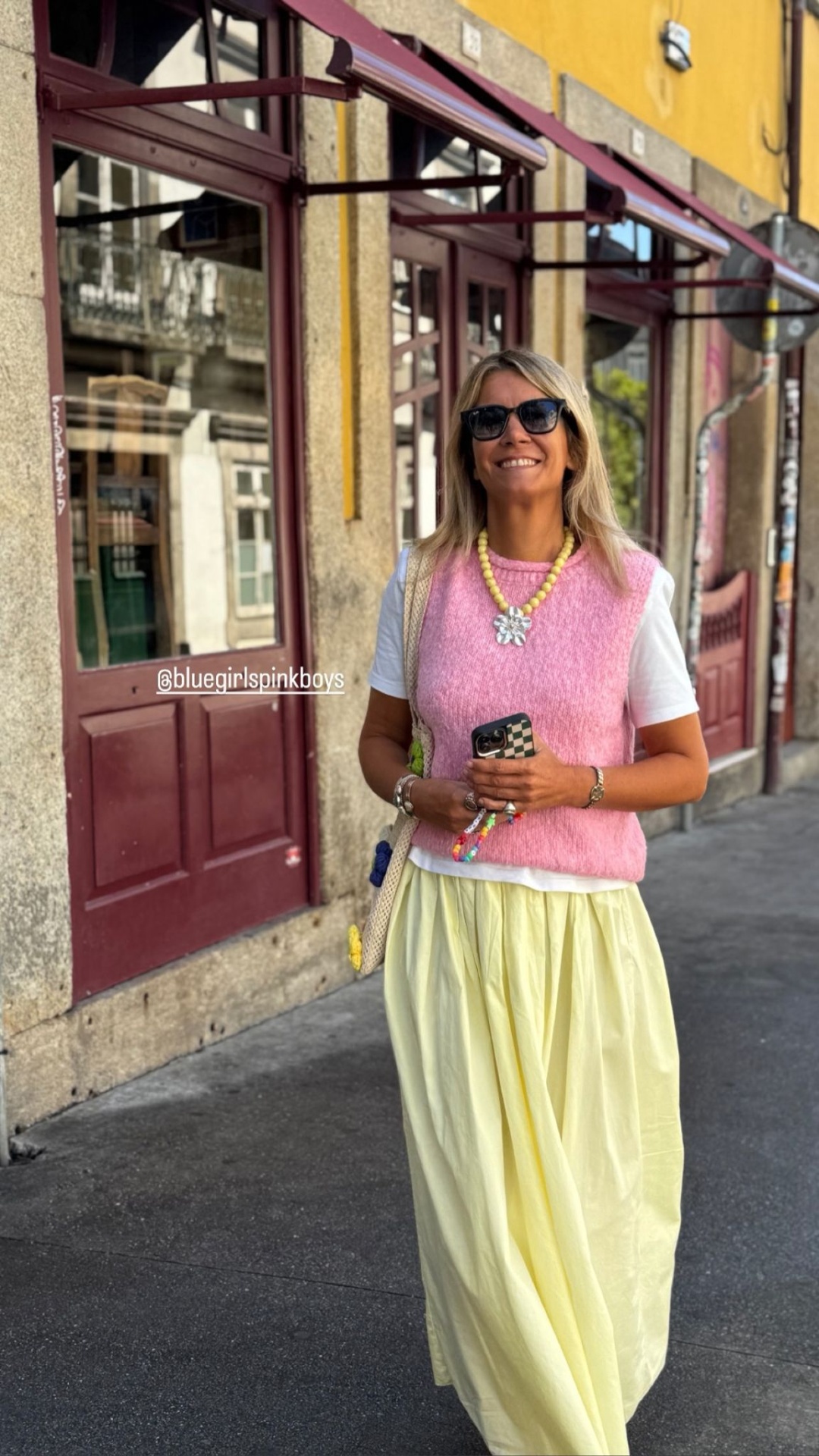 Mulher vestindo colete rosa, saia amarela e t-shirt branca na rua com parede amarela ao fundo
