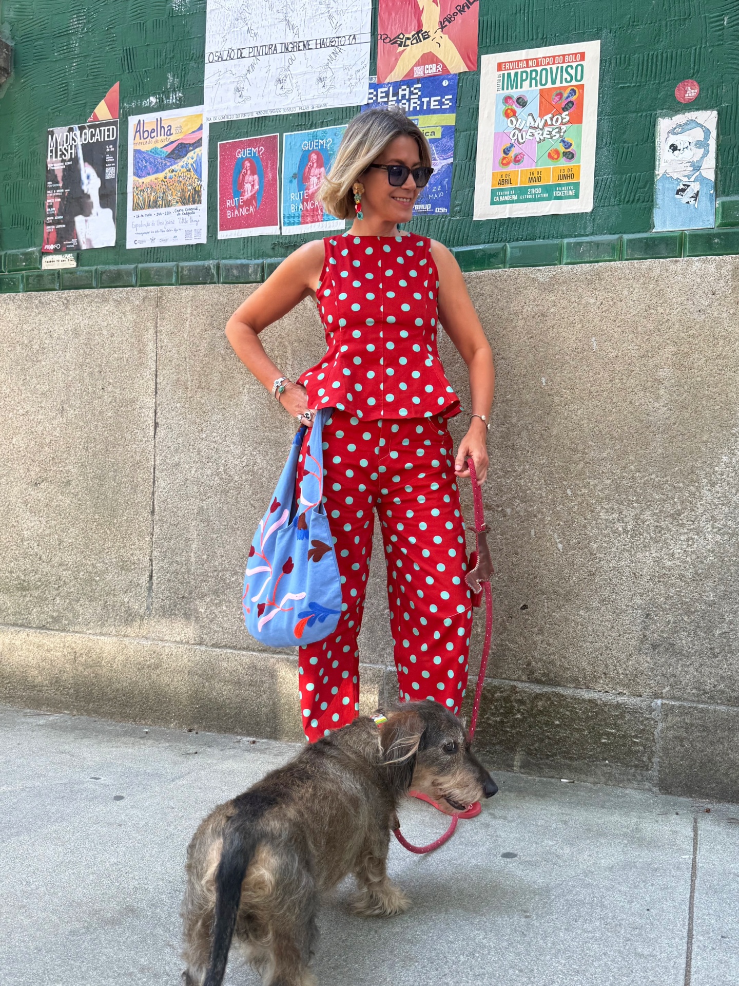 Mulher com conjunto vermelho de bolas azuis e cão com trela vermelha numa rua urbana com cartazes coloridos no muro verde.