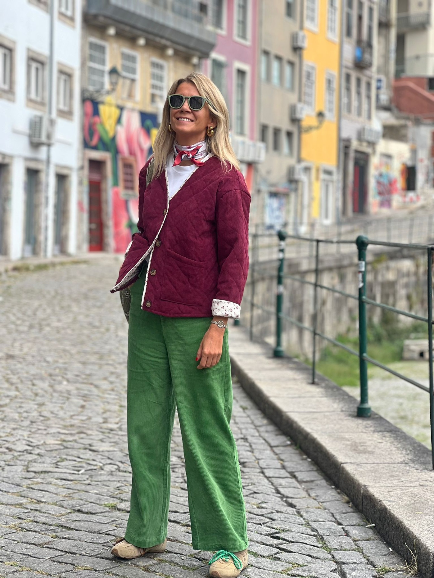 Mulher com casaco borgonha, calças verdes e sapatos beges em rua de calçada