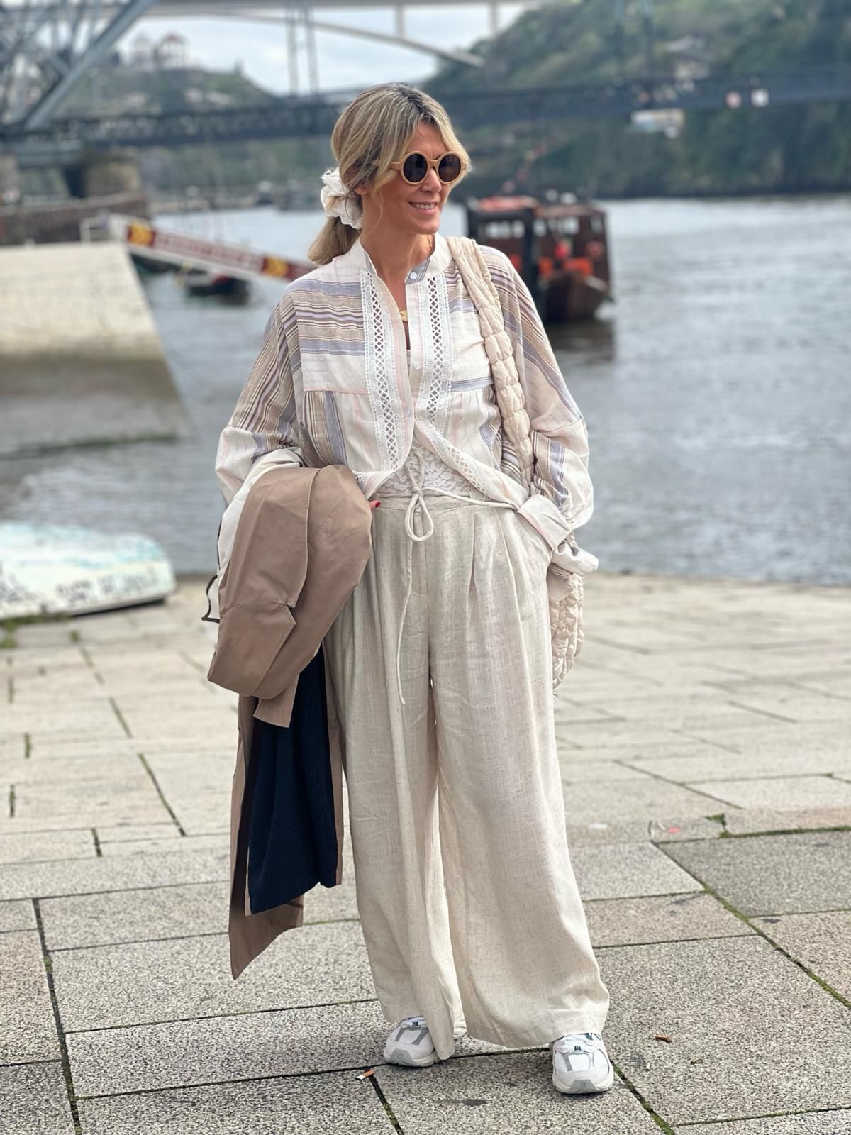 Mulher com roupa casual bege junto a rio com barco e ponte.