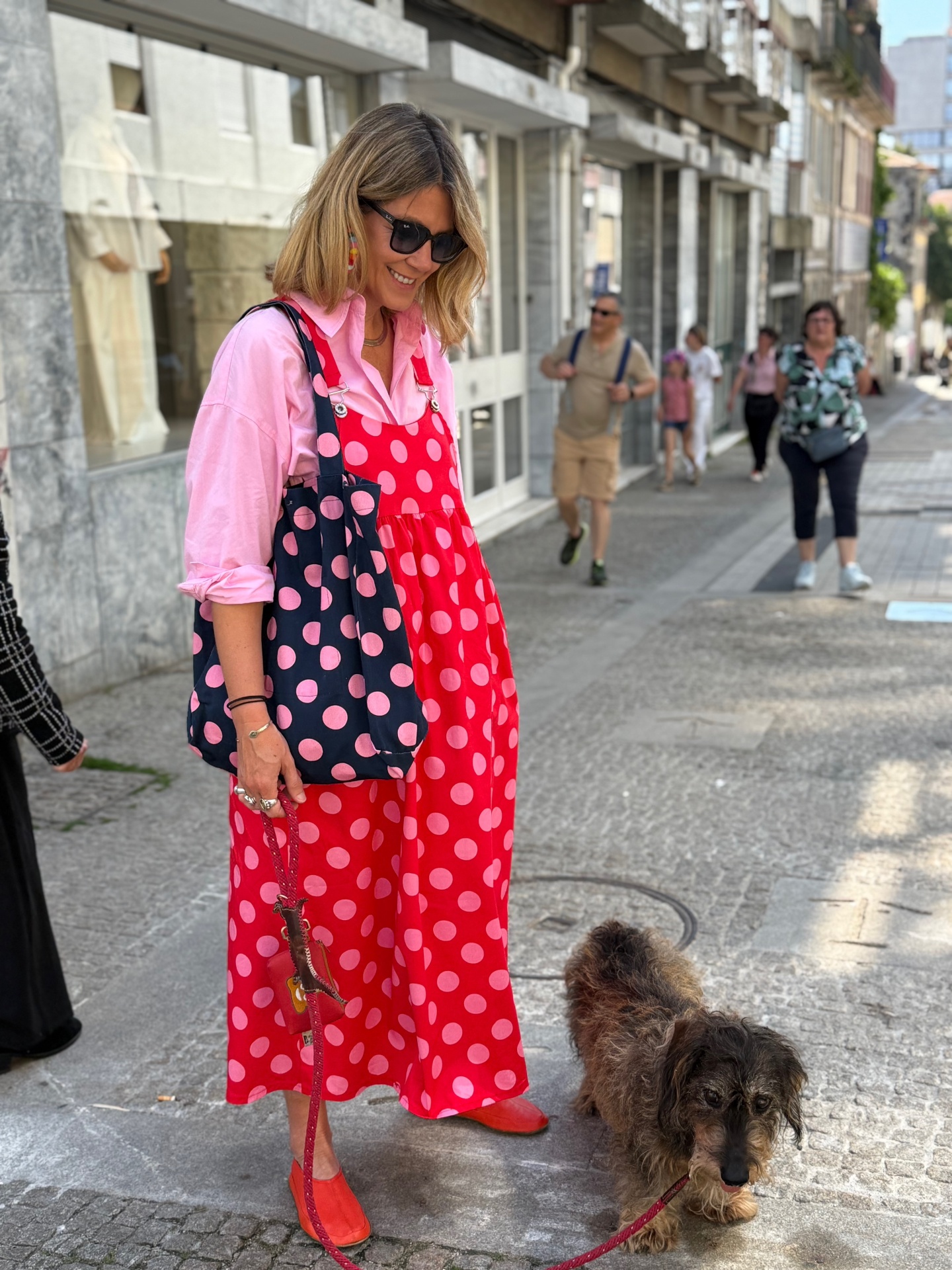Mulher com vestido vermelho de bolas cor-de-rosa e bolsa azul-marinho com bolas cor-de-rosa, com cão castanho na trela