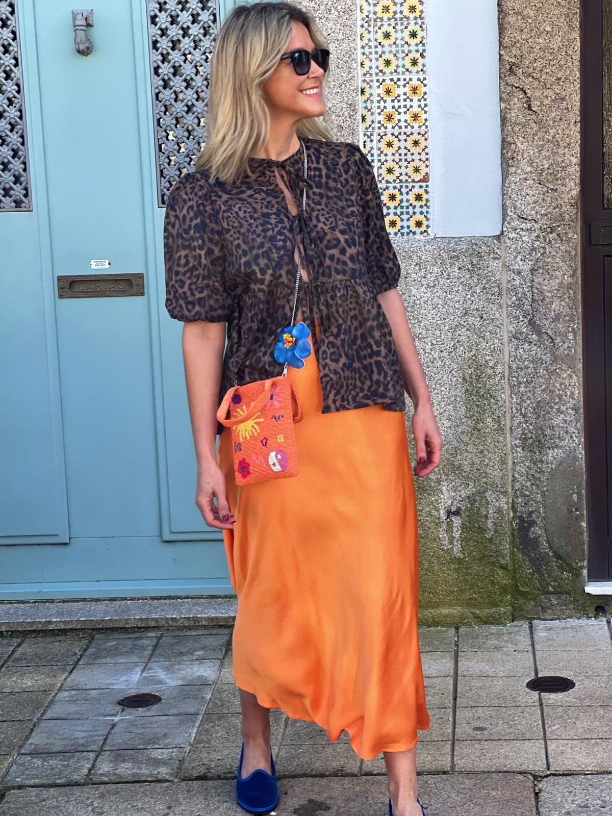 Mulher com blusa animal print, saia laranja, sapatos azuis e bolsa bordada em frente a porta azul com cerâmica