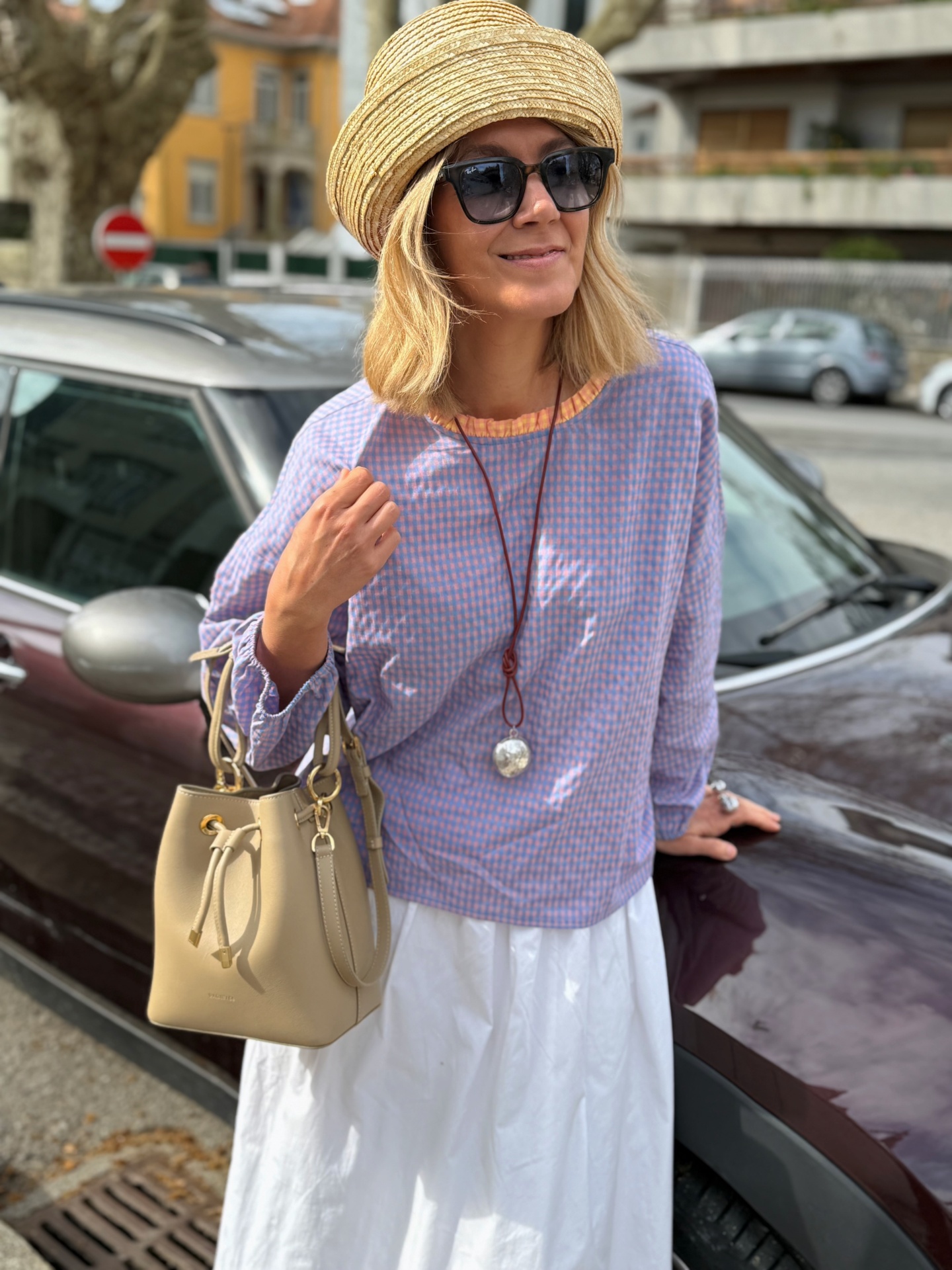 Mulher com chapéu de palha, óculos escuros pretos, blusa xadrez azul e rosa, saia branca, bolsa bege em pele, junto a carro preto na rua.