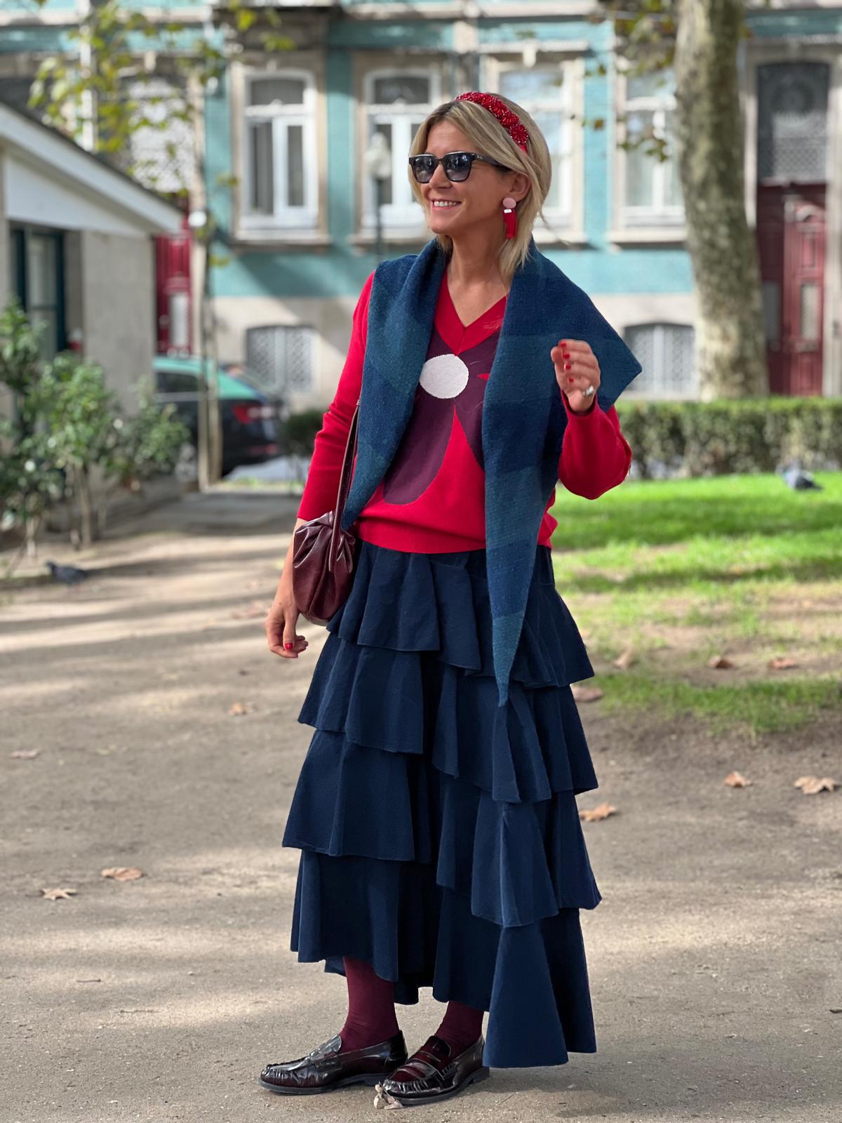 Mulher vestindo saia azul em camadas, camisola vermelha com padrão, lenço azul e óculos de sol