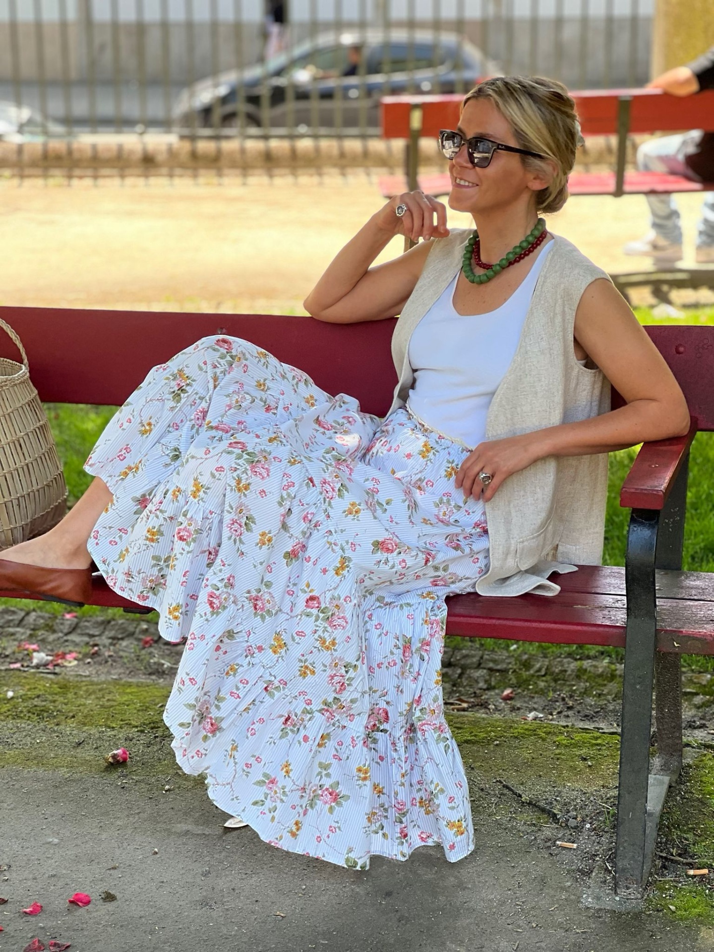 Mulher sentada num banco vermelho usando saia floral, blusa branca e colete bege com óculos de sol.