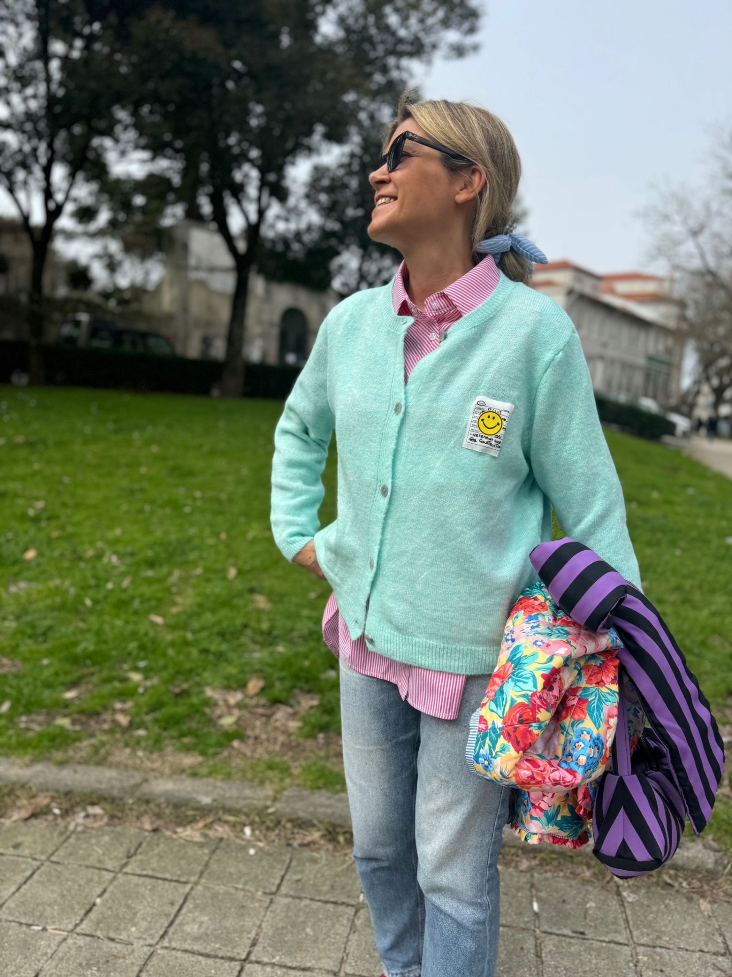 Mulher com cardigan verde claro com patch sorridente, camisa rosa, jeans e lenço azul segurando casaco colorido em parque