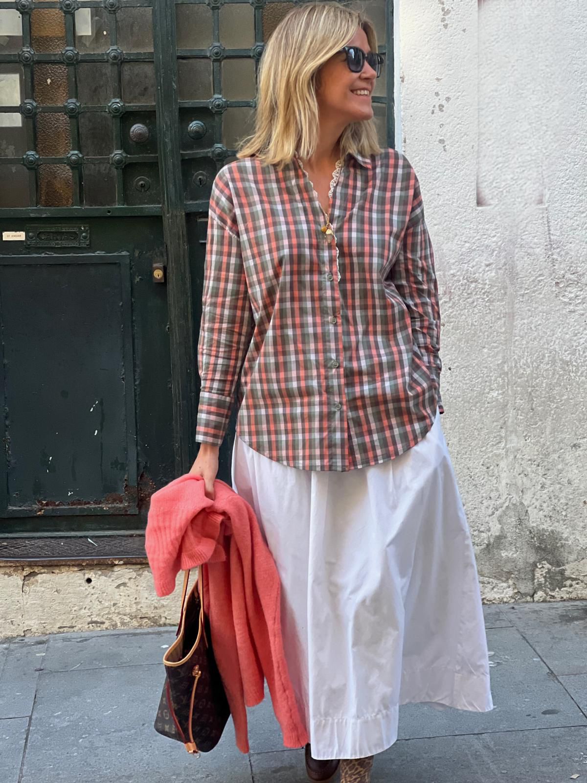 Pessoa com camisa xadrez castanha e rosa e saia branca, segurando casaco coral e mala castanha
