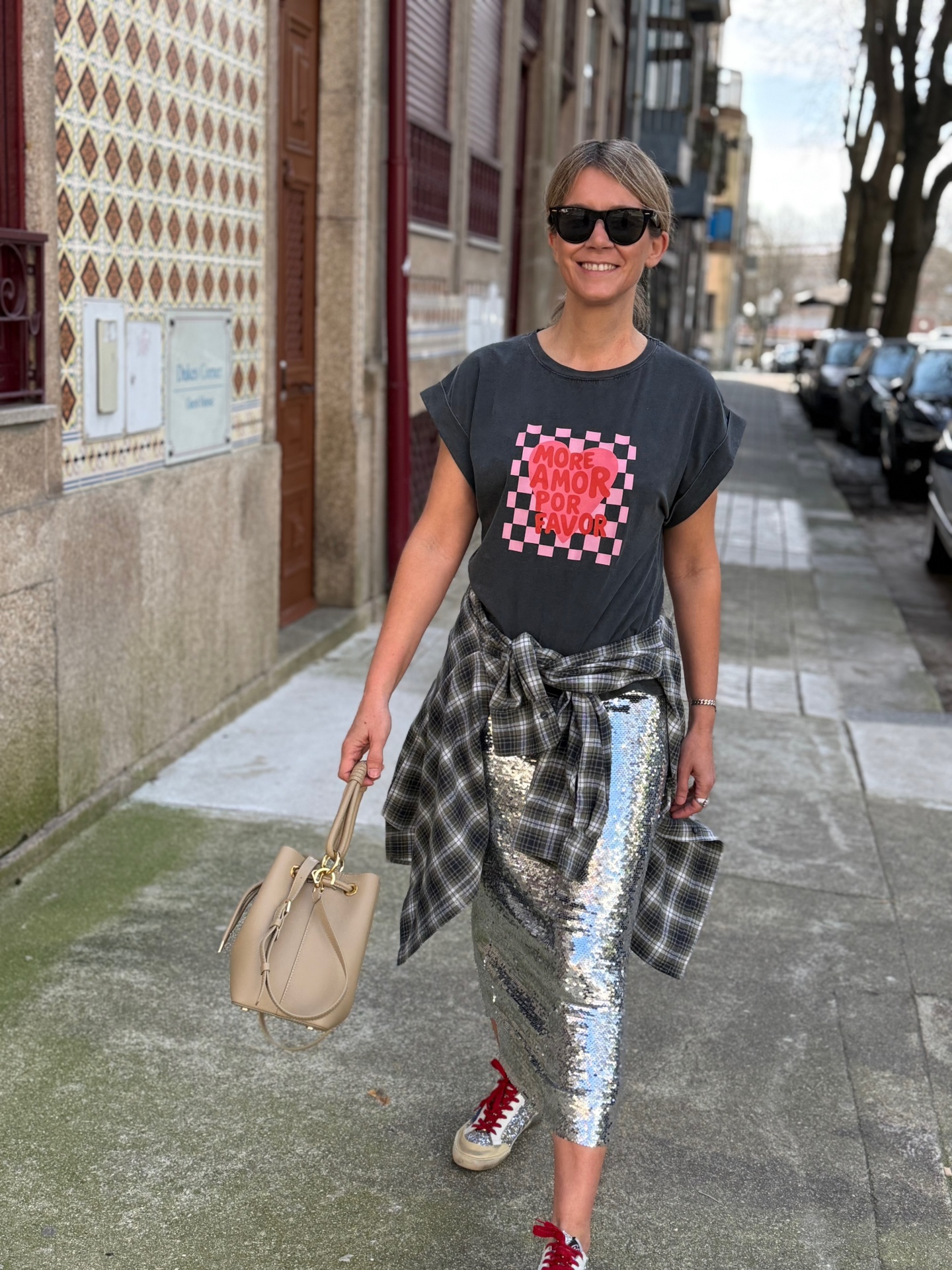 Mulher com t-shirt preta com texto vermelho, saia de lantejoulas prateada, camisa xadrez amarrada na cintura e mala bege