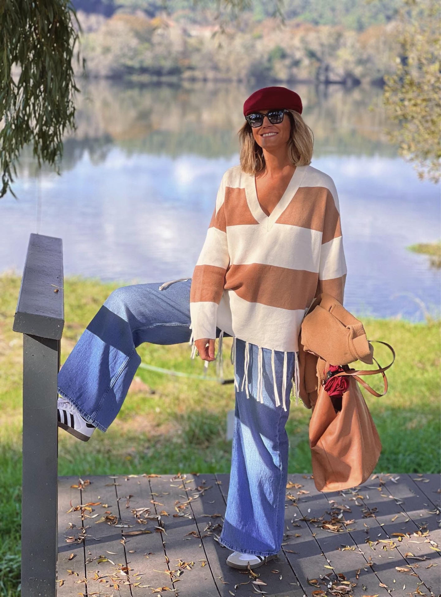 Mulher com casaco de malha riscas beges e brancas, calças jeans largas, ténis brancos, óculos escuros e boina vermelha junto a lago com árvores