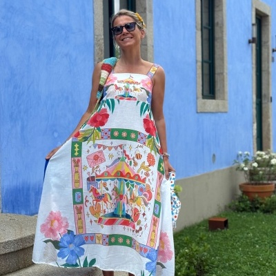 Mulher com vestido longo estampado colorido ao ar livre junto a parede azul e janelas.