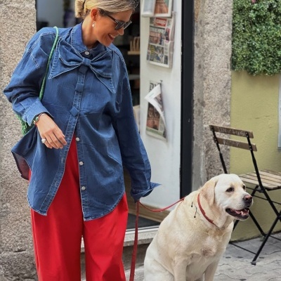 Mulher com camisa azul de denim com laço, calças vermelhas, óculos escuros e cão labrador creme sentado
