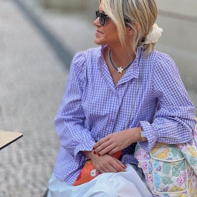 Mulher usando blusa xadrez lilás e branco, saia branca, e bolsa colorida patchwork