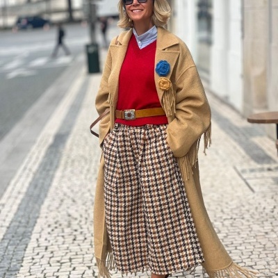 Mulher com casaco bege, camisola vermelha, saia pied-de-poule, chapéu azul, óculos escuros e sapatos vermelhos numa calçada urbana