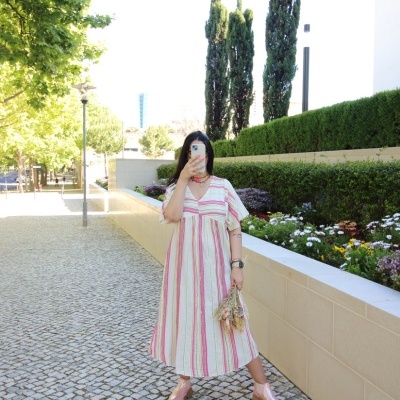 Mulher com vestido de algodão às riscas verticais rosa e branco, sapatos rosa, segurando flores secas numa rua com calçada portuguesa e vegetação
