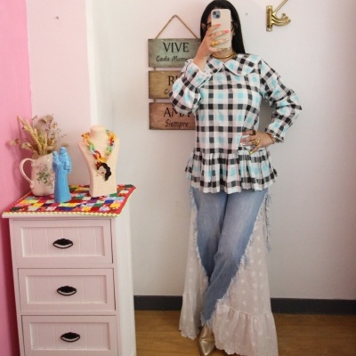 Pessoa com blusa xadrez, calças de ganga e sapatos dourados, tirando selfie junto a cómoda e placa na parede.