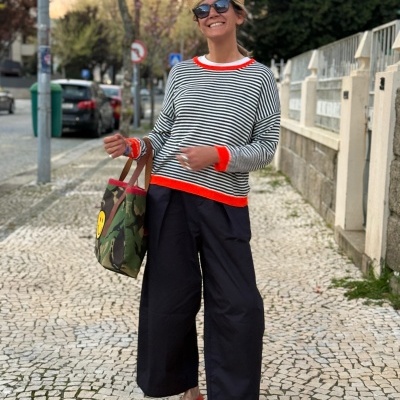 Mulher com camisola riscada, calças largas, sapatos vermelhos e bolsa camuflada numa calçada.