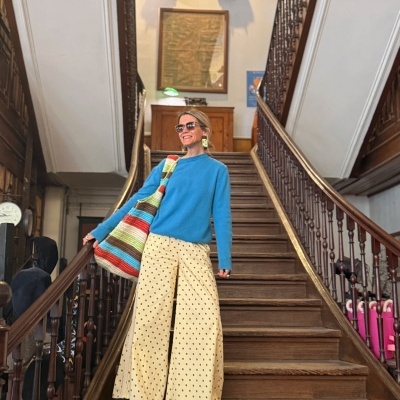 Mulher com camisola azul, calças largas bege com bolinhas e saco colorido numa escadaria em madeira