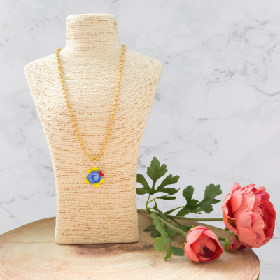 Colar dourado com pendente colorido em busto de tecido e flores rosas ao lado