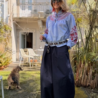 Pessoa com blusa azul clara com riscas brancas e bordados vermelhos, calças pretas largas, cinto de couro, sapatos com tachas, óculos escuros e brincos vermelhos, segurando copo de vinho