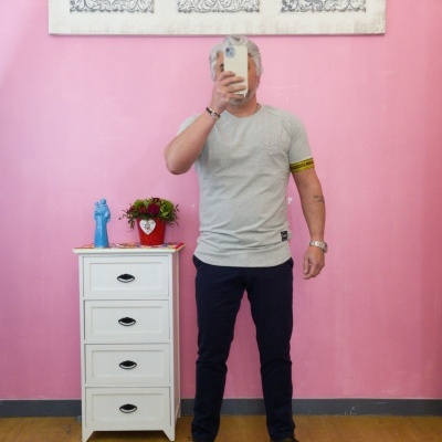 Homem com t-shirt cinza e calças escuras a tirar foto em quarto com parede rosa e móvel branco