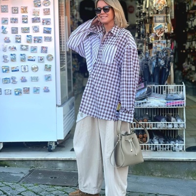 Mulher com camisa xadrez castanha e branca, calças creme largas, mocassins e mala bege na rua com lojas ao fundo
