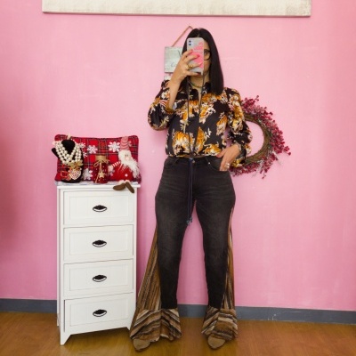 Pessoa vestindo blusa estampada e calças com franjas diante de parede rosa e móvel branco com acessórios