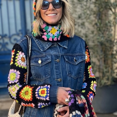 Mulher com casaco de ganga azul com mangas e capuz em crochet colorido, usando óculos escuros e segurando tecido de crochet