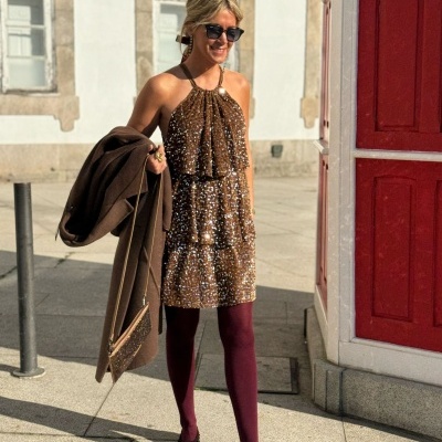 Mulher de vestido curto castanho com padrão e collants bordô segura casaco castanho em rua urbana