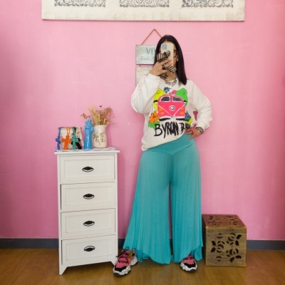 Pessoa a usar camisola branca estampada e calças largas azuis, de pé contra parede rosa com móvel branco ao lado