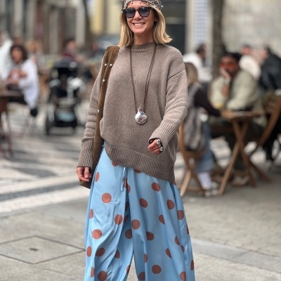 Mulher com camisola castanha, calças azuis com bolinhas castanhas e lenço na cabeça numa rua com esplanada.