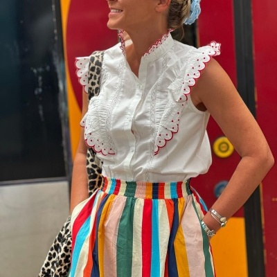 Mulher com blusa branca bordada, saia listrada colorida e óculos escuros em fundo colorido