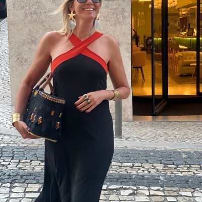 Mulher com vestido longo preto e vermelho, acessórios dourados e mala preta na calçada