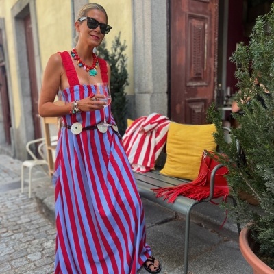 Mulher com vestido riscado azul e vermelho, colar colorido e óculos escuros segurando copo junto a banco com almofada amarela e manta às riscas