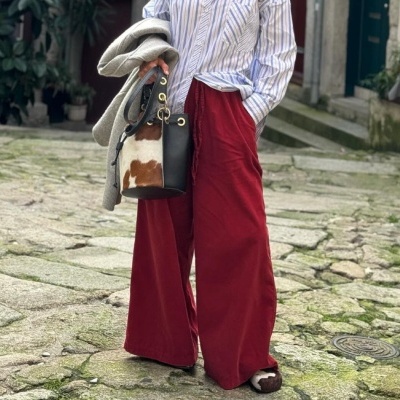 Pessoa com calças largas vinho, camisa de riscas, sapatos e bolsa com padrão pele de vaca, casaco cinzento, em rua de pedra