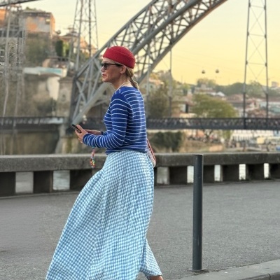 Mulher com camisola azul às riscas brancas, saia azul e branca padrão vichy, sapatos vermelhos e chapéu vermelho numa avenida junto a ponte metálica.