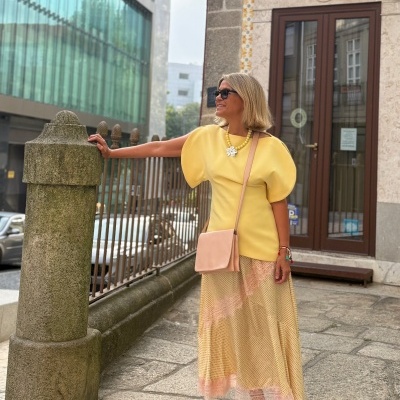Mulher de vestido amarelo e saia comprida bege com acessórios coloridos em ambiente urbano.