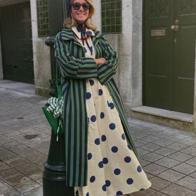 Mulher com casaco riscado verde e preto, vestido creme com bolinhas azuis, sapatos bege, saco verde com lenço, gorro azul e óculos escuros