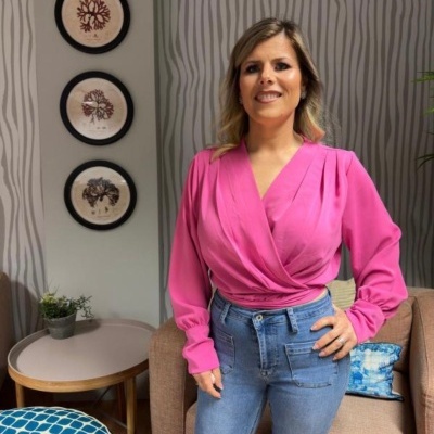 Mulher com blusa rosa e calças de ganga azuis em sala decorada com sofá, planta e quadros arbóreos
