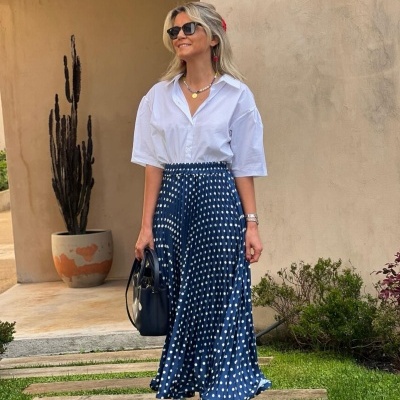 Mulher com camisa branca, saia azul de bolinhas, sapatos vermelhos e óculos de sol junto a parede bege