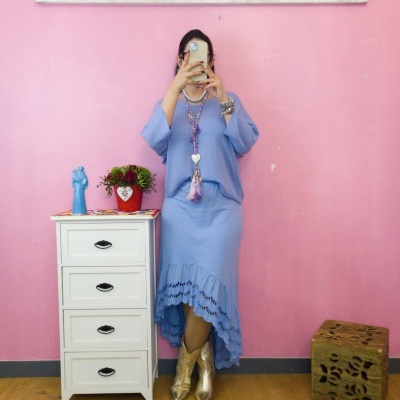 Pessoa com vestido azul claro e botas douradas encostada em cómoda branca com decoração, parede rosa e quadros decorativos.