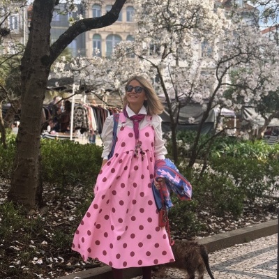 Mulher com vestido rosa às bolinhas castanhas na rua à sombra de árvores floridas com cão e calçada de mosaico.