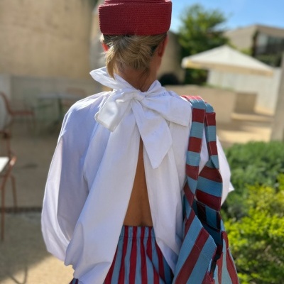 Pessoa com camisa branca aberta nas costas e saia riscada vermelha e azul, chapéu vermelho e bolsa riscada vermelha e azul, vista de costas