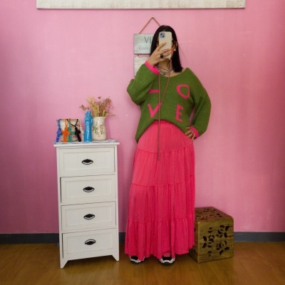 Mulher a tirar selfie com suéter verde e saia rosa em quarto com parede e móveis decorativos