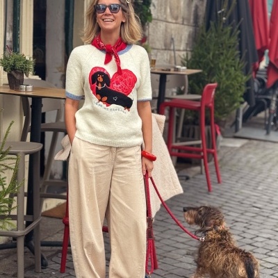 Mulher com camisola branca bordada, calças creme, mocassins castanhos e lenço vermelho, com cão na trela vermelha numa calçada urbana